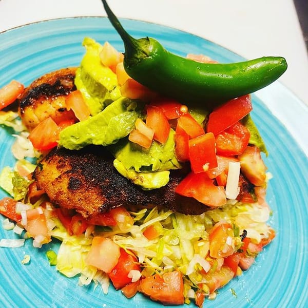Mexican dish with grilled chicken, avocado, diced tomatoes, lettuce, and a green chili on a blue plate.