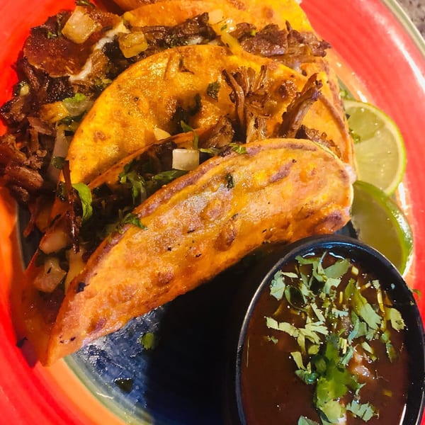 Three golden-fried El Camino Real birria tacos with limes and a side of consommé on a colorful plate.